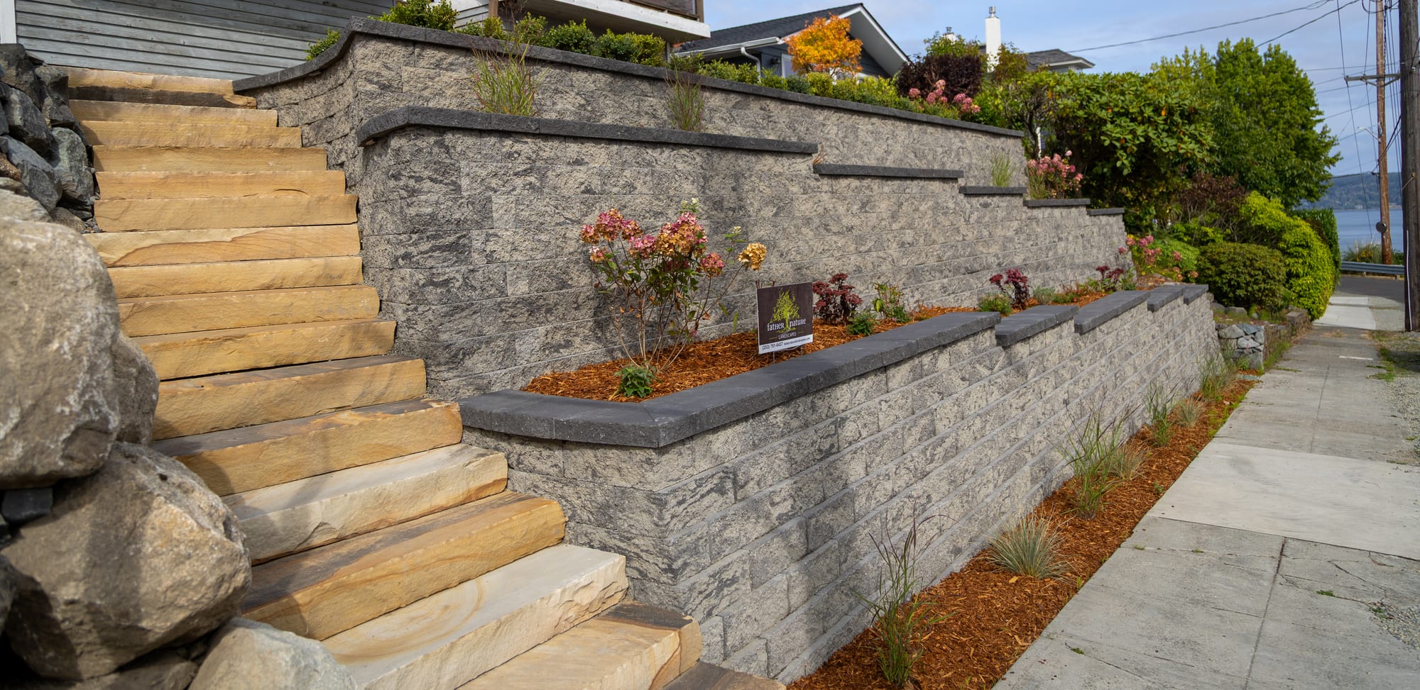 Stone slab steps climbing beside tiered block retaining walls with charcoal caps and planting terraces, a Father Nature Landscapes yard sign in the bed, and water in the distance