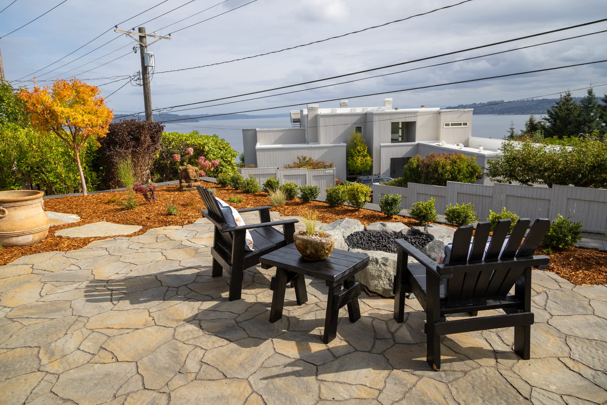 Two Adirondack chairs and a low table beside the boulder fire pit on the paver patio, with a view over the neighborhood to the water