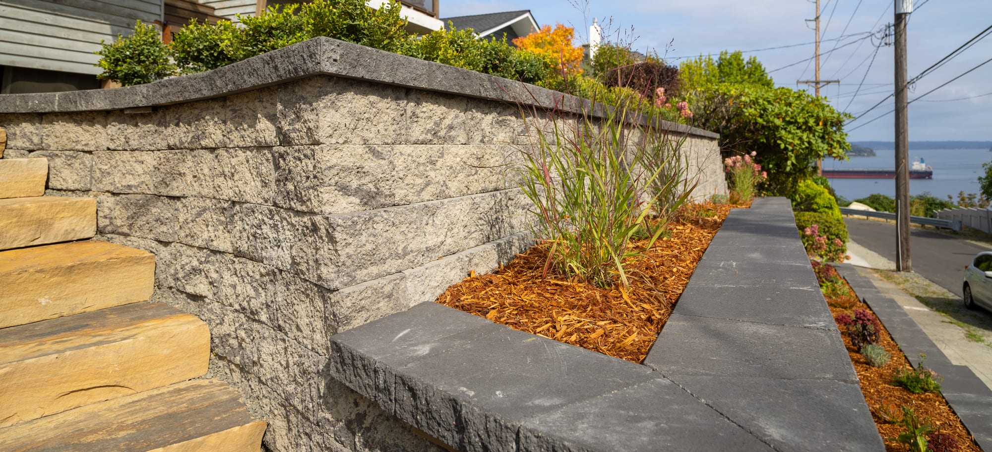 A retaining wall corner with a dark charcoal cap and ornamental grass in the planting terrace, with the sidewalk and water beyond